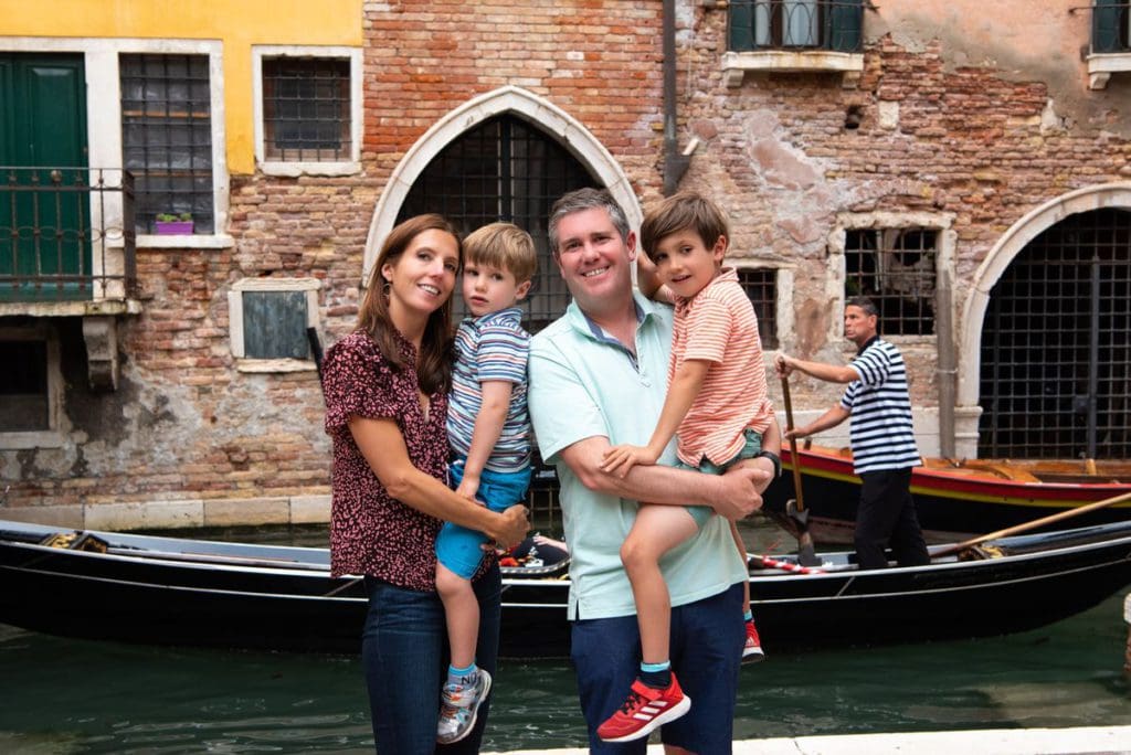 A family of four stands together with a Venetian canal and gondola behind them in Venice, Italy, one of the top places in travel in 2023 with kids.