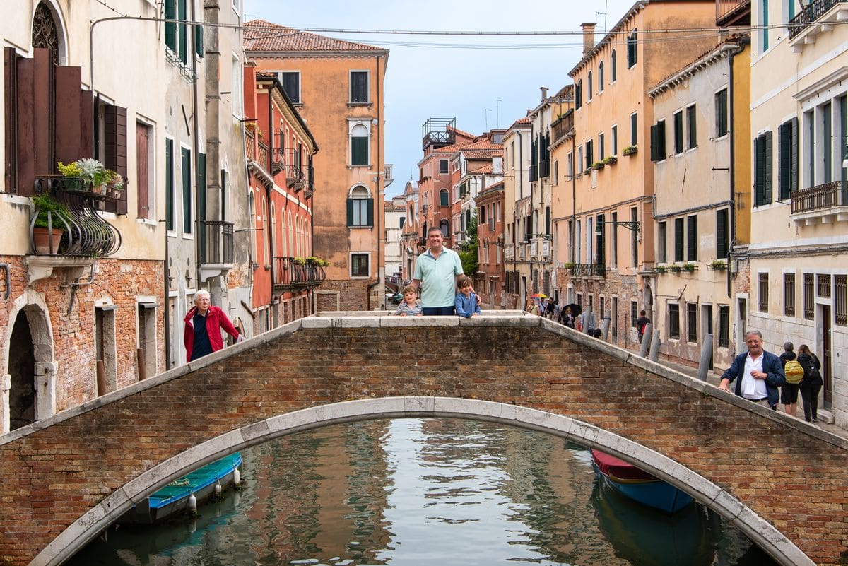 A dad and his two young sons stand together on a bridge near Canareggio in Venice with the canal extending into the distance behind them.