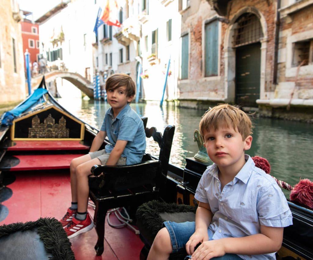 Two young boys ride in a gondola down a canal in Venice, Venetian buildings behind them. A gondola ride is one of the best things to include on a Venice itinerary with kids. 