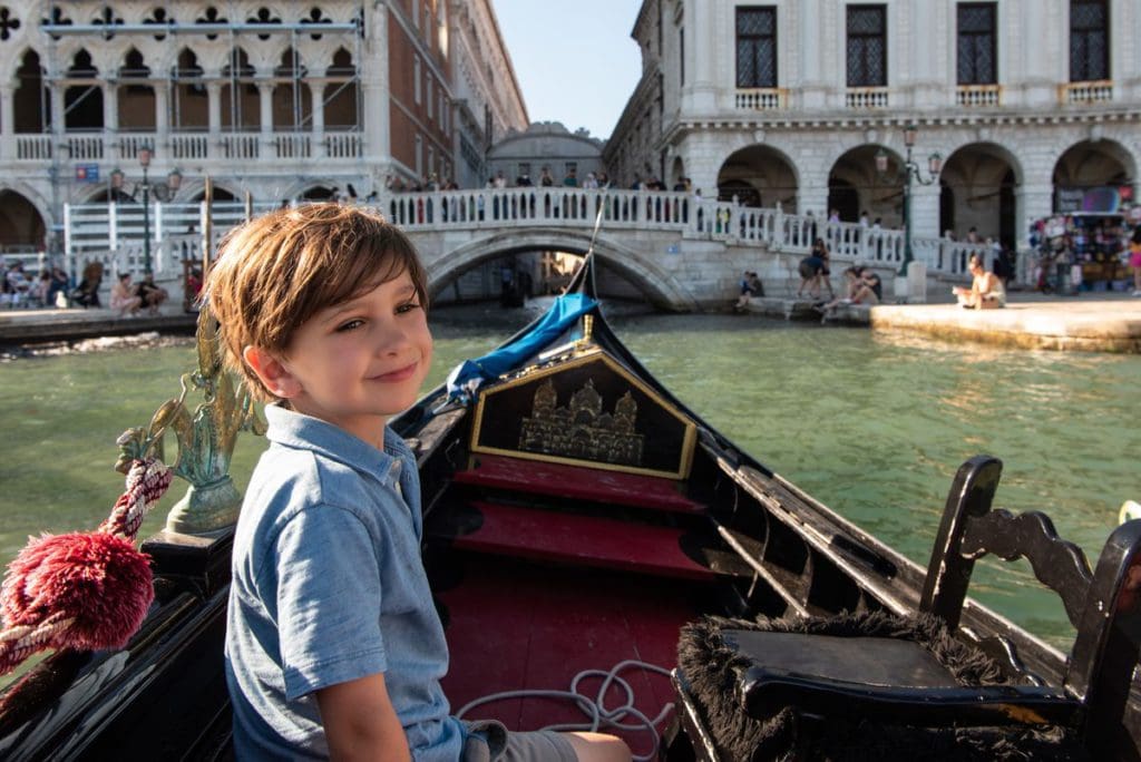 A young boy smiles as he rides a gondola through the canals of Venice, with the Bridge of Sighs in the distance.