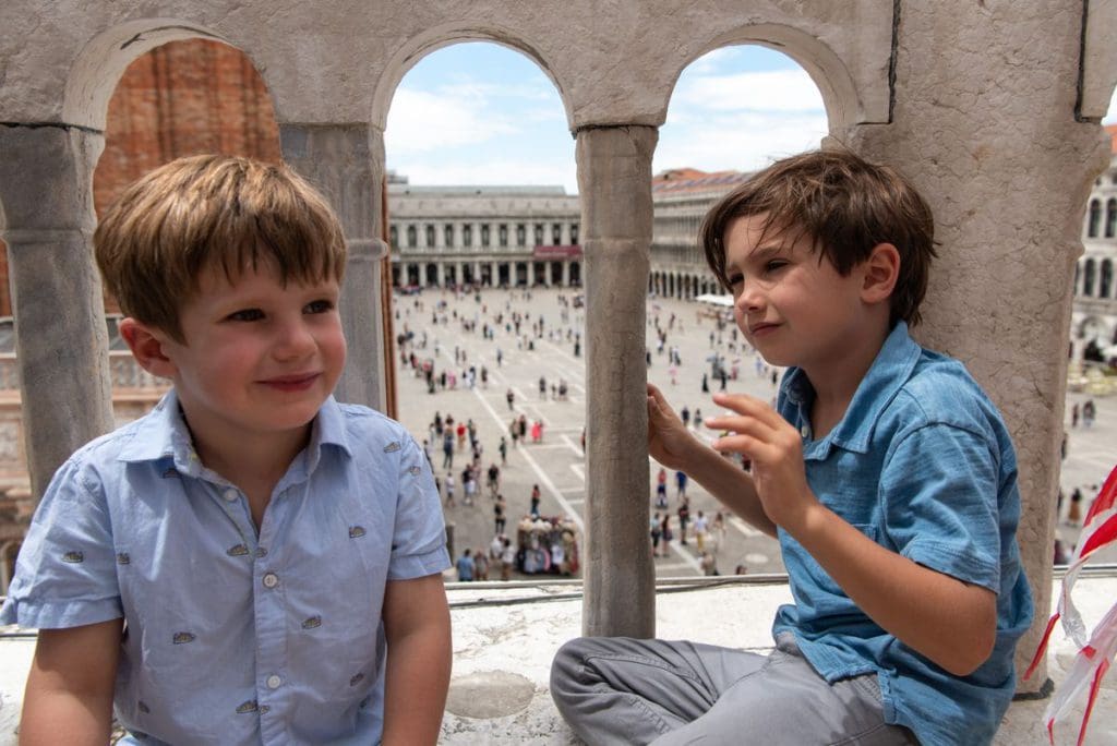 Two boys look through slats in the stone of St. Mark's Basilica down to San Marco Piazza.