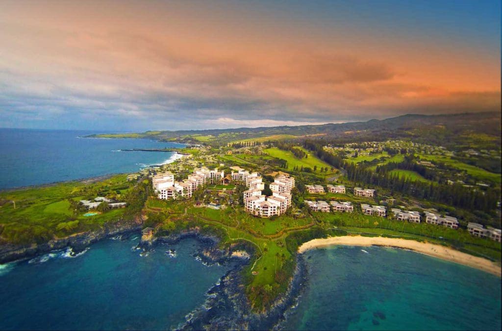 An aerial view of the Montage Kapalua Bay at sunset, featuring an oceanfront property and large resort buidlings.