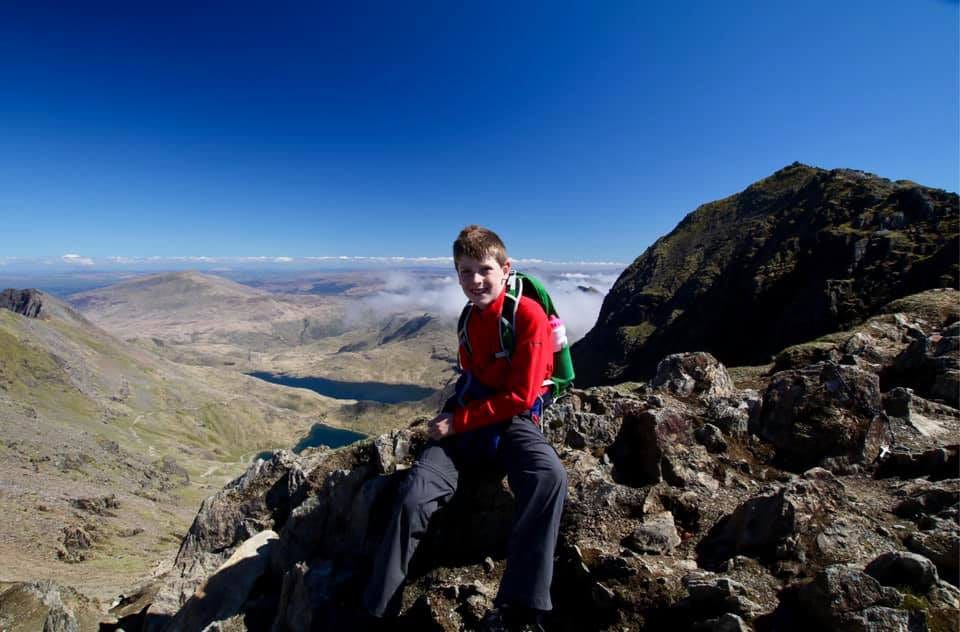 A young boy sits on a large rock while hiking in Wales, with an expansive view of the Welsh countryside behind him.