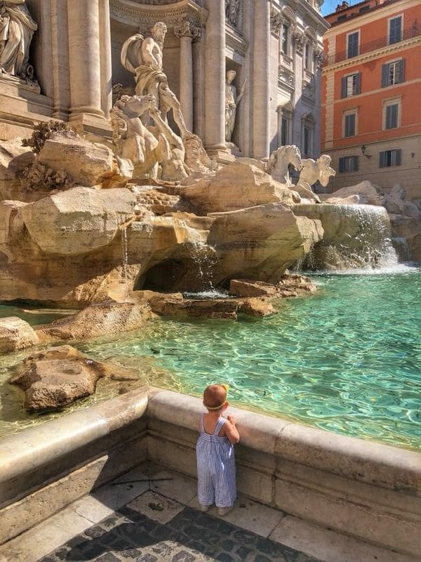 An infant girl stands at the edge of the Trevi Fountain looking over the ledge into the water.