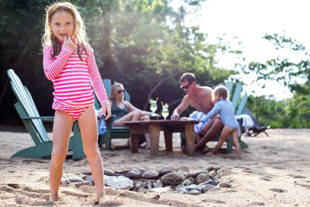 A young girl smiles at the camera, while her family enjoys drinks around a beach table at Sweet Songs Jungle Lodge, a Muy’Ono Resort.