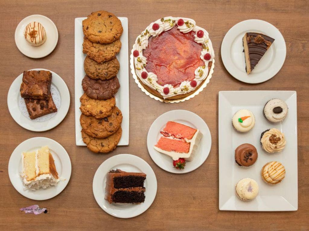 A table filled with treats from The CakeRoom Bakery, including cookies, cakes, cupcakes, and pie.