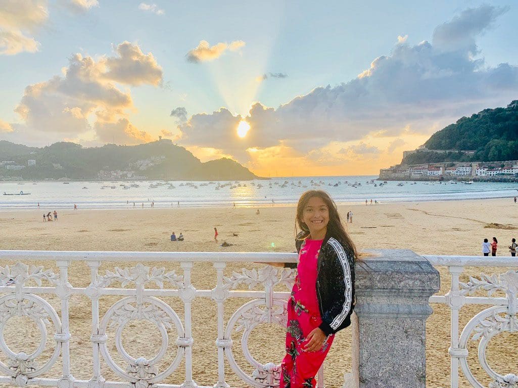 A young girl stands along a fence with the beach at Donostia-San Sebastian behind her, one of the best mild weather European destinations for a family summer vacation.