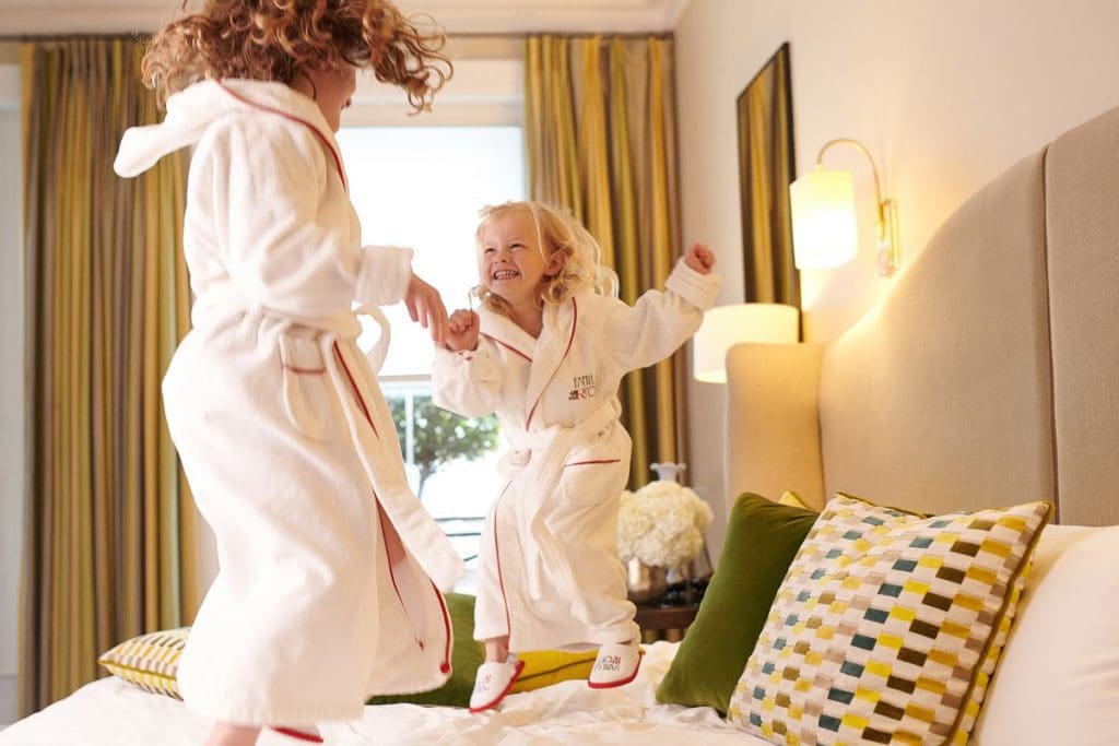 A mom and her young daughter jump on a hotel bed in their room at the Brown's Hotel, a Rocco Forte Hotel, while wearing hotel robes at one of the best London Hotels for Families.