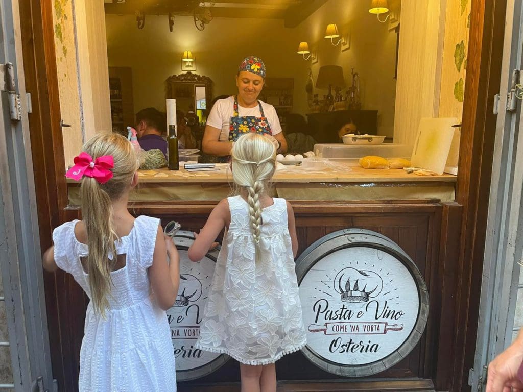 Two young girls watch a woman make pasta through a window at Pasta e Vino Osteria in Trastevere, one of the best neighborhoods to stay in Rome with kids.