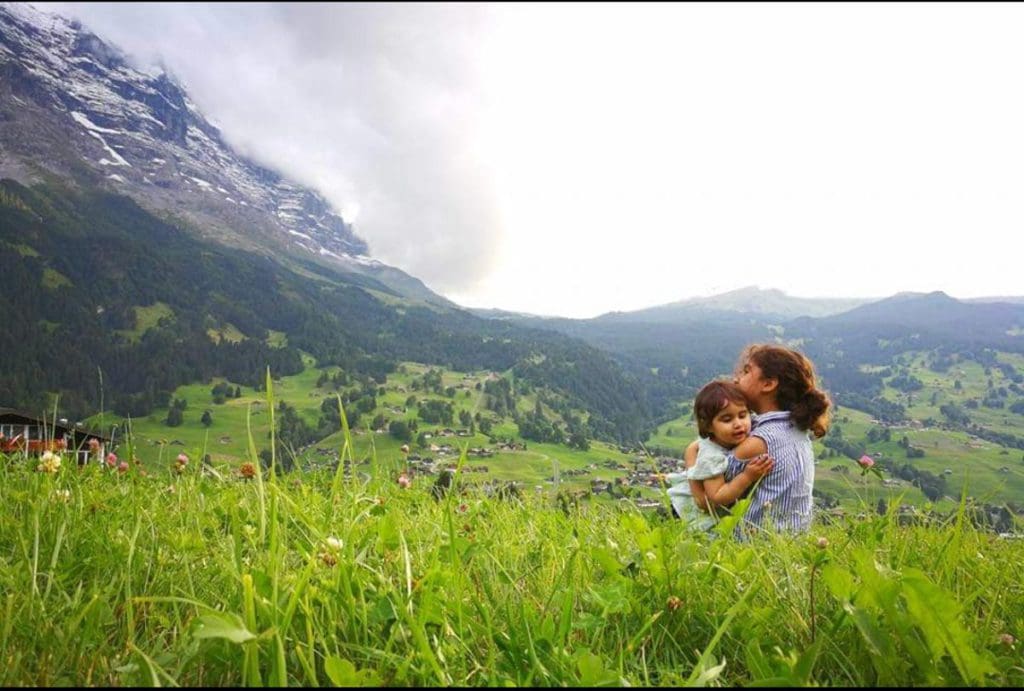 Two small kids embrace while sitting in a green pasture near Grindelwald, with a mountain view near one of the best towns and villages to visit with your family in Switzerland.