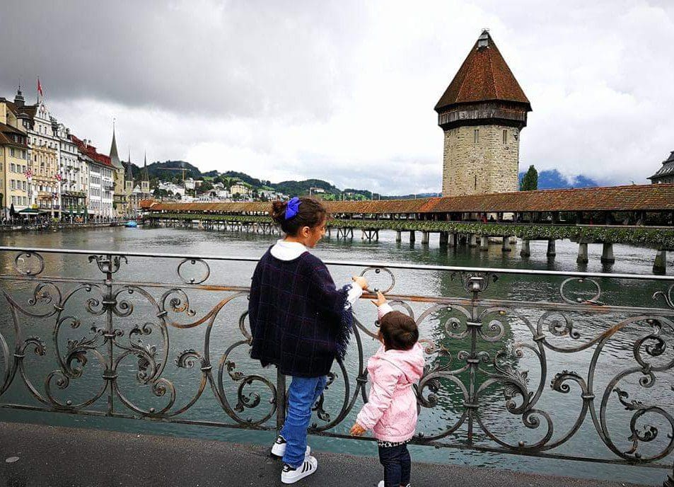 Two girls stand along a fence looking at the iconic lake and bridge in Lucerne, one of the best towns and villages to visit with your family in Switzerland.