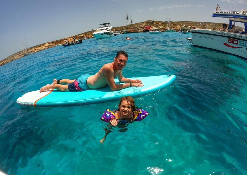 A dad lays on a SUP, while paddling next to his daughter swimming in the water off the coast of Malta.