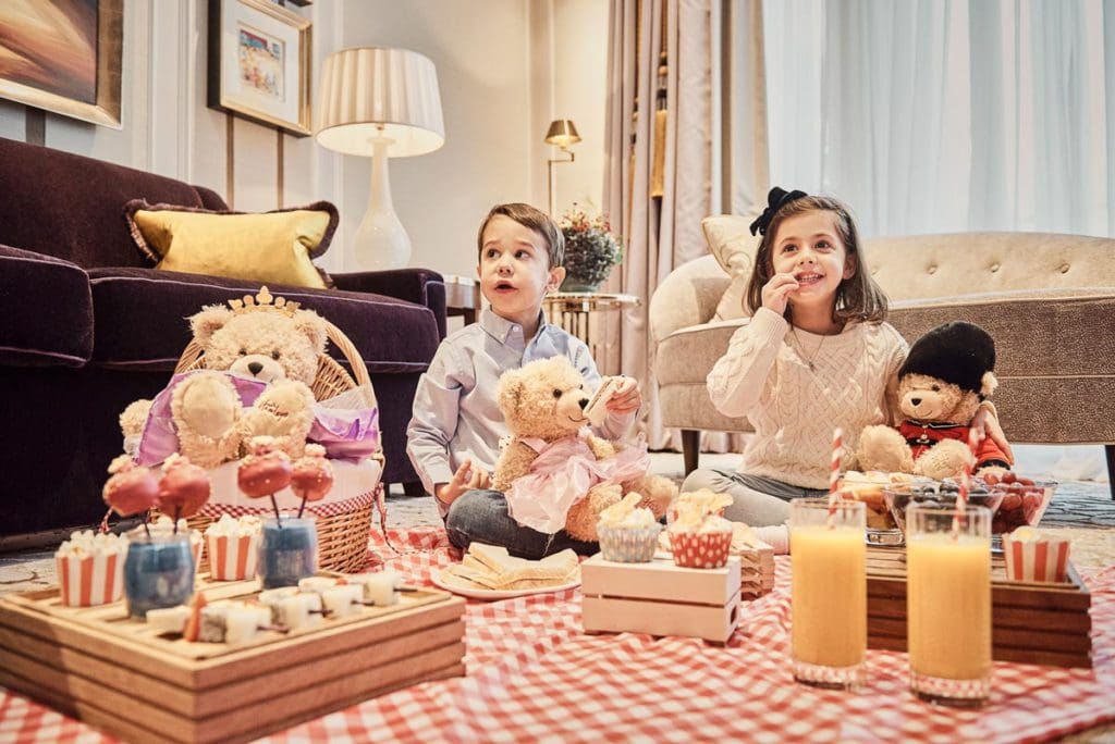 Two kids sit inside a room at London Marriott Hotel Park, with many toy bears and sweets.