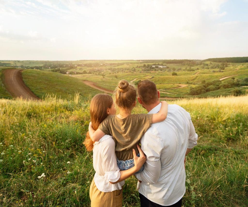 A mom, dad, and their young daughter stand together enjoy a lovely green field view.