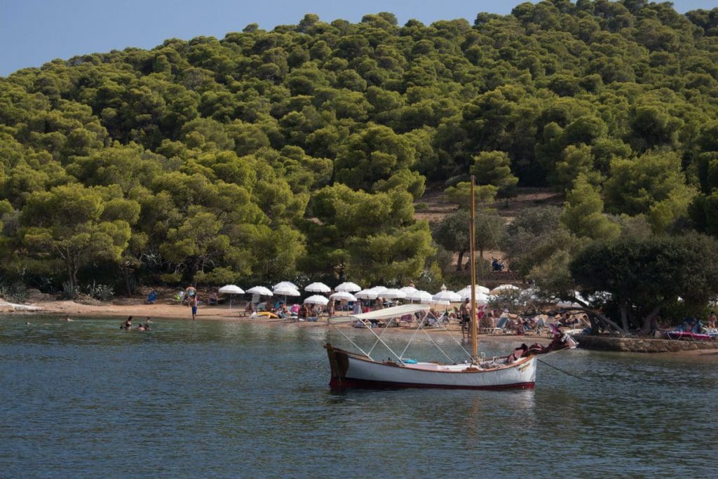 A boat rocks gently in the water, off-shore from Spetses, a Greek island, with cabanas on shore and a dense wooded area in the distance.