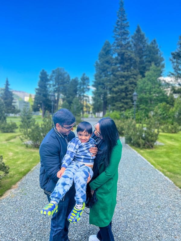 A family of three poses together, smiling, on a paved path in St. Moritz with tall trees in the distance.