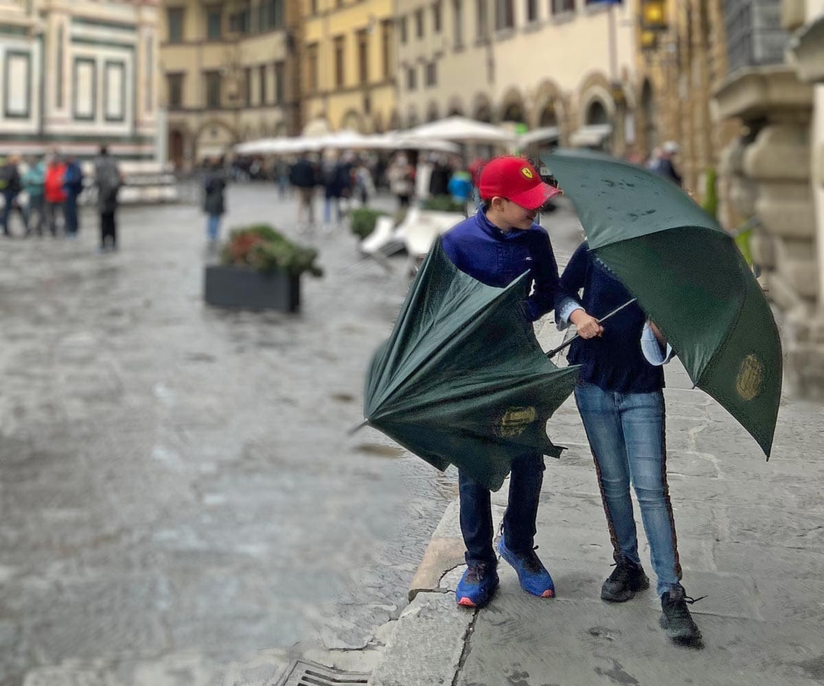 Two kids walk along a street in Florence with umbrellas on a rainy day in Italy, one of the top places in travel in 2023 with kids.