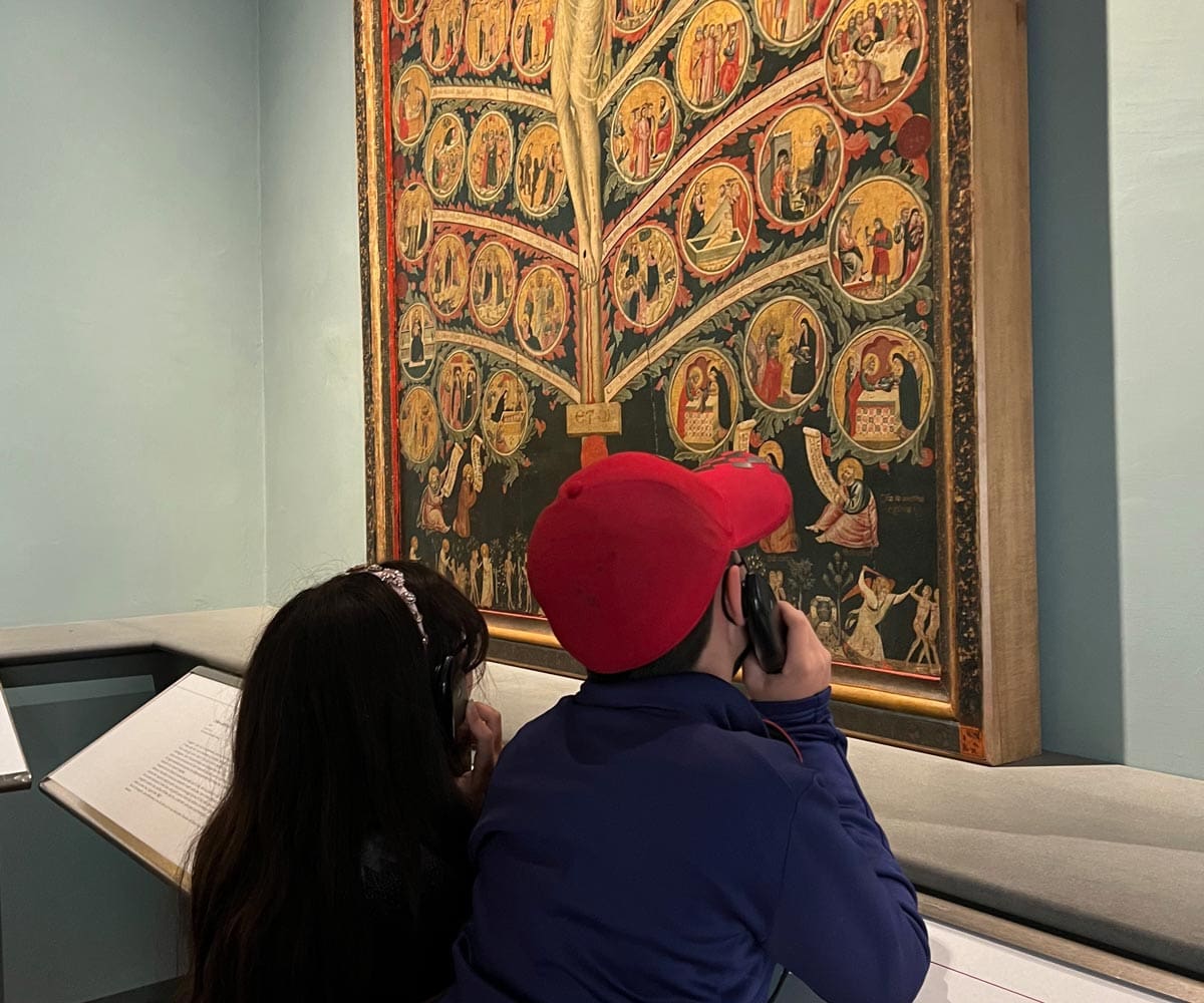 Two kids, listening to an audio tour at a Florence museum, look up at a large painting.