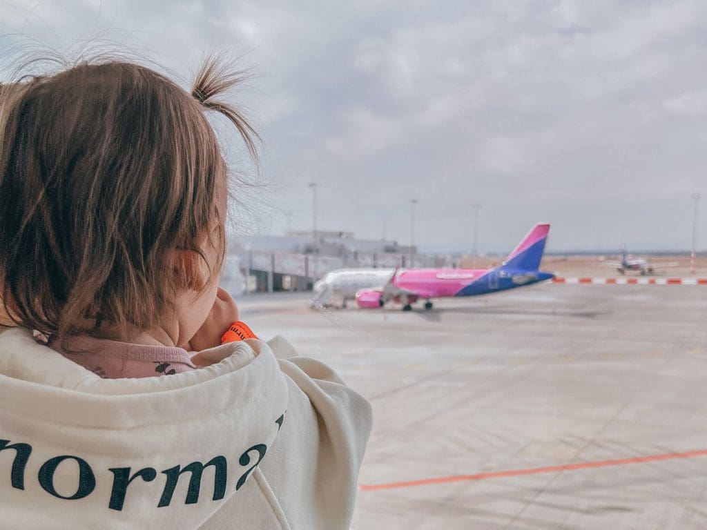 A toddler girl looks out an airport window toward an airplane on the tarmack.