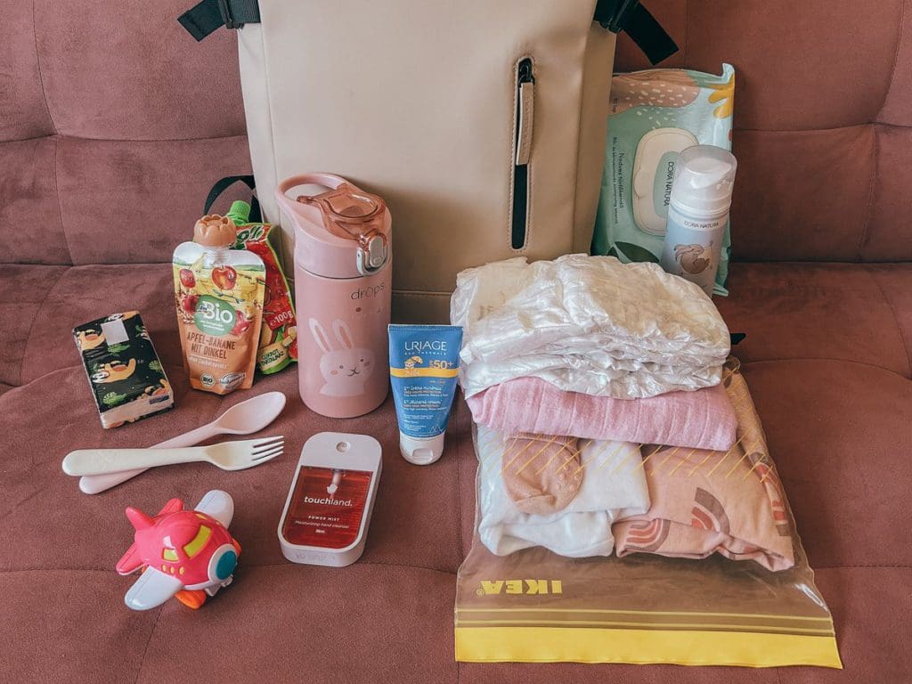 Several items laid out, ready to be packed in a backpack for a trip, including diapers, toy, food, and wipes, which are all included on our is a must-pack list for a European vacation with toddlers.