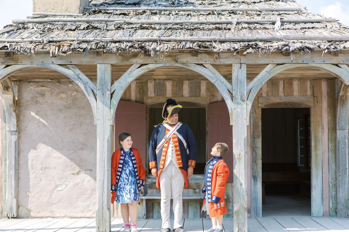 Two kids stand with a costumed reenact-or at Fort Michilimackinac in Upper Michigan, one of the best affordable summer vacations in the United States with kids.