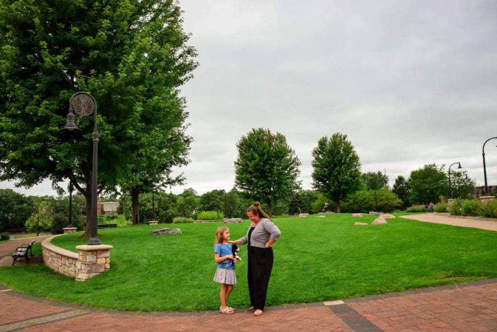 A young girl and her mom stand on a verdant lawn with trees in the distance at Phoenix Park.