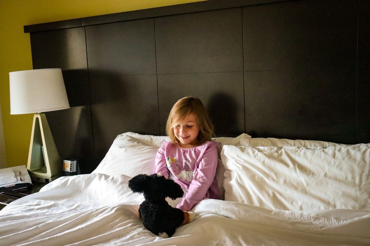 A young girl sits on a large bed with her stuffie, while staying at the Staybridge Suites Eau Claire - Altoona.