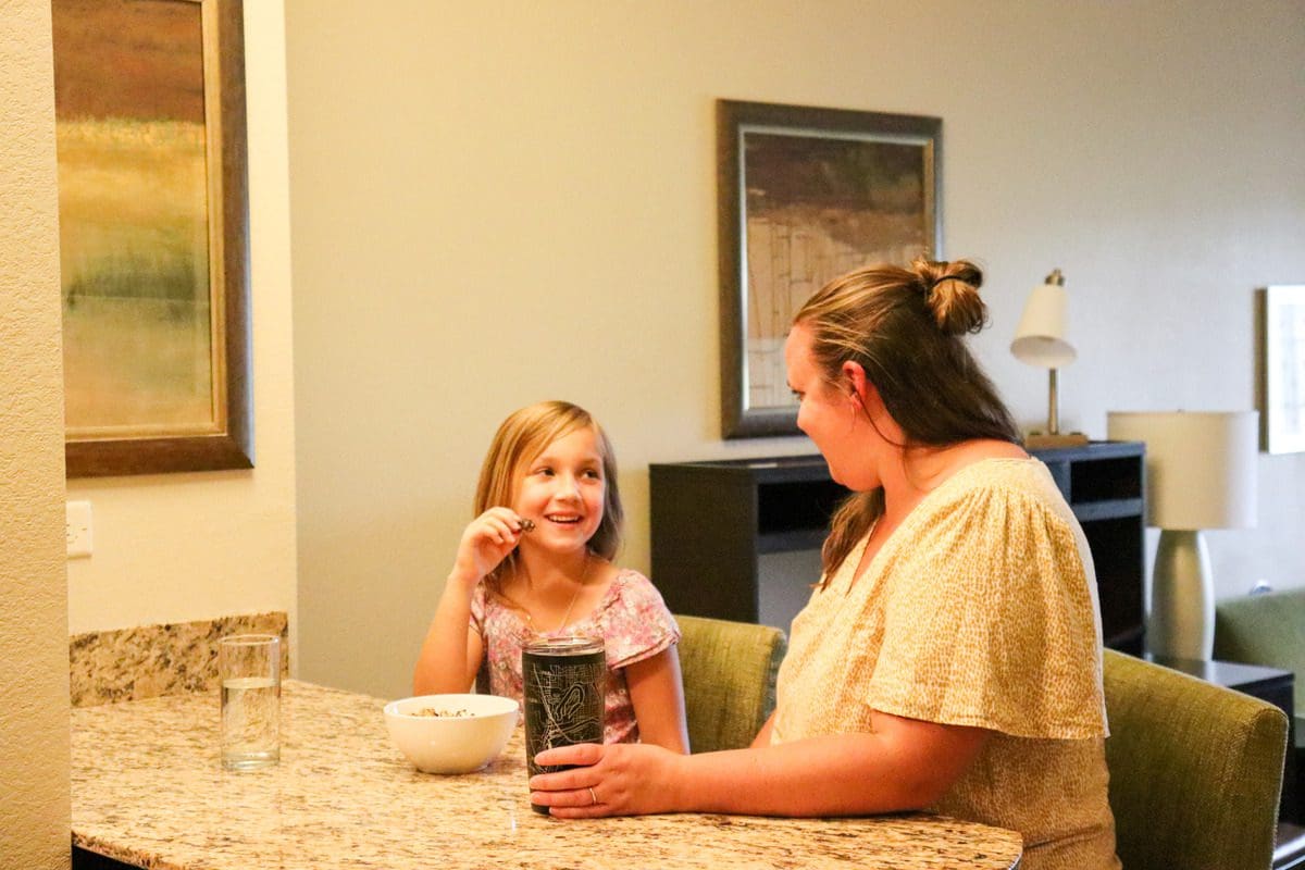 A mom and her young daughter sit at the counter of their kitchen in a hotel suite, while staying at the Staybridge Suites Eau Claire - Altoona.