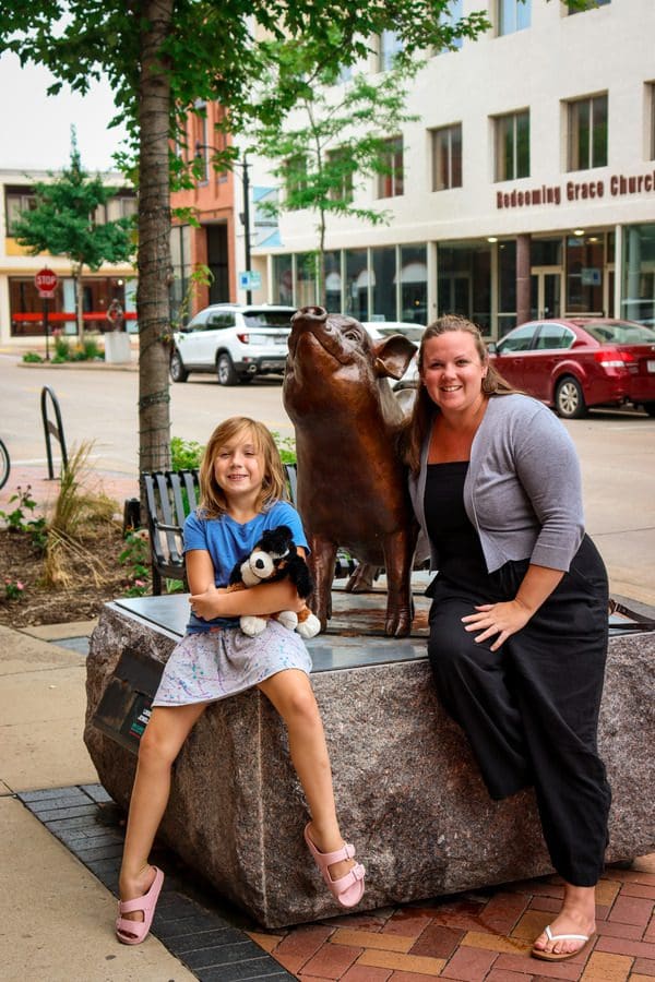 A mom and her young daughter smile as they pose with a large smiling pig sculpture on the Eau Claire Sculpture Tour, one of the best things to do in Eau Claire with kids.