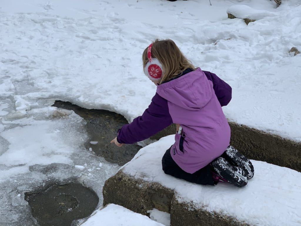 A young girl kneels in the snow, while pointing at an open area in the flowing river at Nerstrand Big Woods State Park.