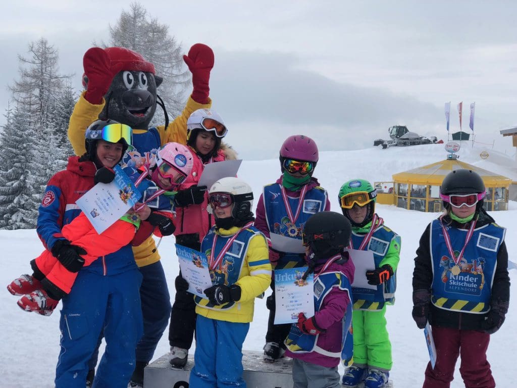 Kids excitedly stand with their medals and the ski mascot after their final race during ski school, a fun addition to this family skiing in Alpendorf itinerary.