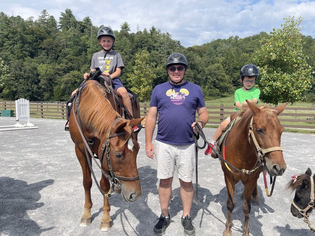 A dad stands between two horses, atop which his two kids sit respectively, while exploring the grounds of Blackberry Farm.