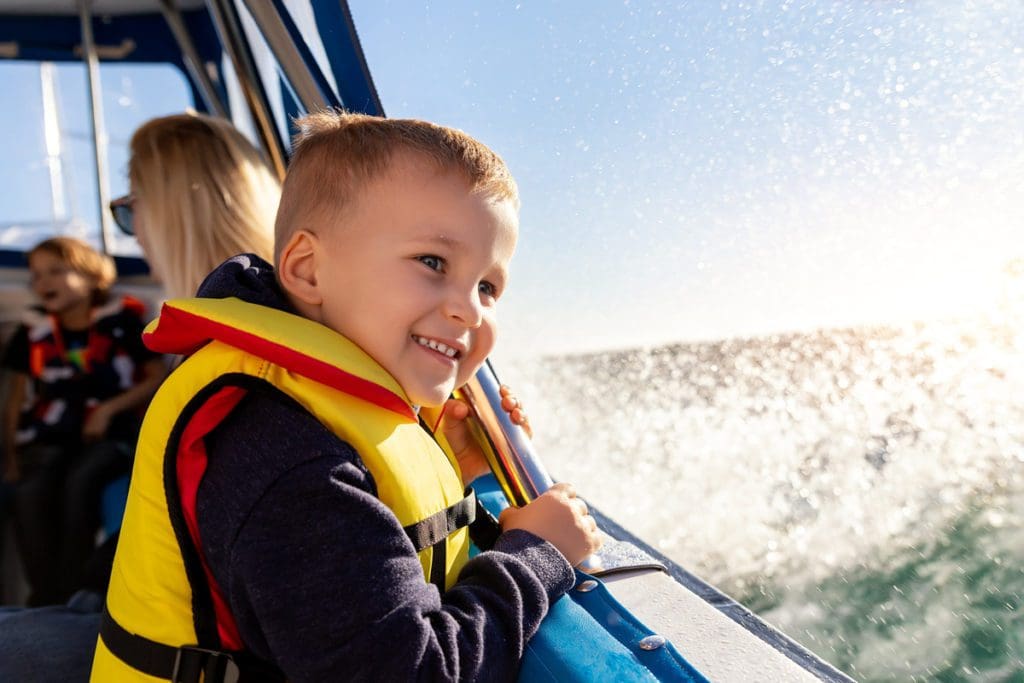 A little boy wearing a life jacket smiles broadly while waves splash onside of the sail boat.