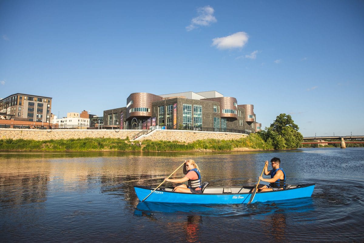 Two people canoe along a river in Eau Claire, with downtown in the distance, on a summer day.