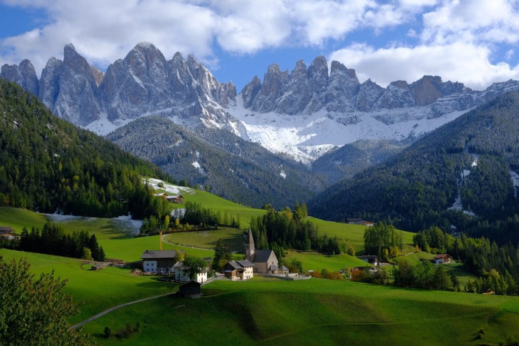 A view of a verdant valley and small building structures with the Dolomites in the distance.