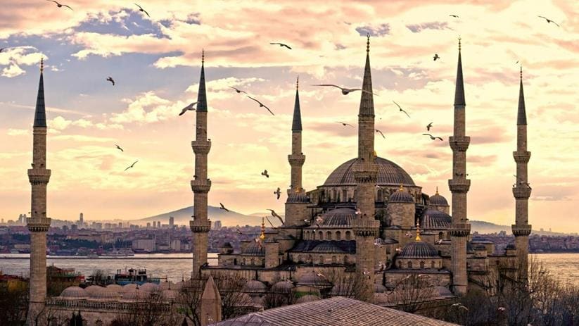 The exterior of the Blue Mosque at dusk, with seagulls flying around.