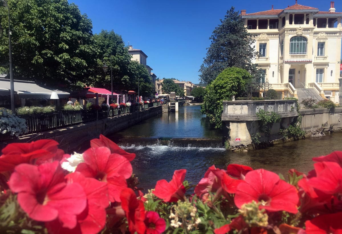 A lovely view of a canal in L'Isle-Sur-la-Sorgue through a row of red flowers on a lovely day in France.