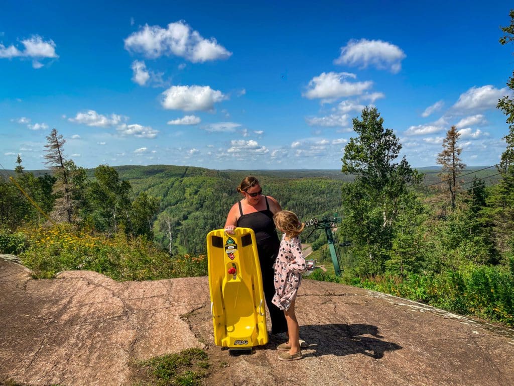 A mom and her daughter stand together with an alpine slide and a sweeping view of Lutsen Mountain behind them, one of the best places to visit in northern Minnesota with kids.