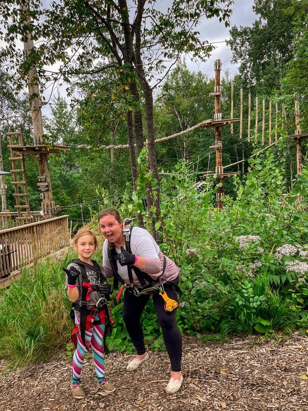 A mom and her daughter stand together, both wearing ropes course harnesses, while zip-lining at the North Shore Adventure Park.