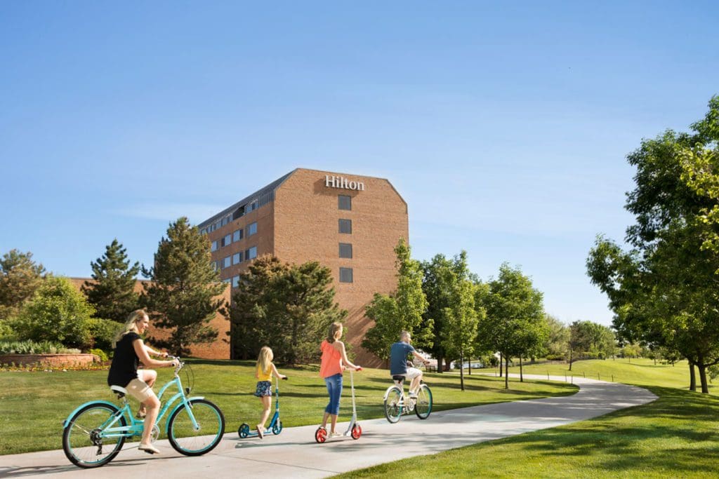 A family of four, riding respective bikes and scooters enjoys a sunny day down a path that passes the Hilton Denver Inverness.