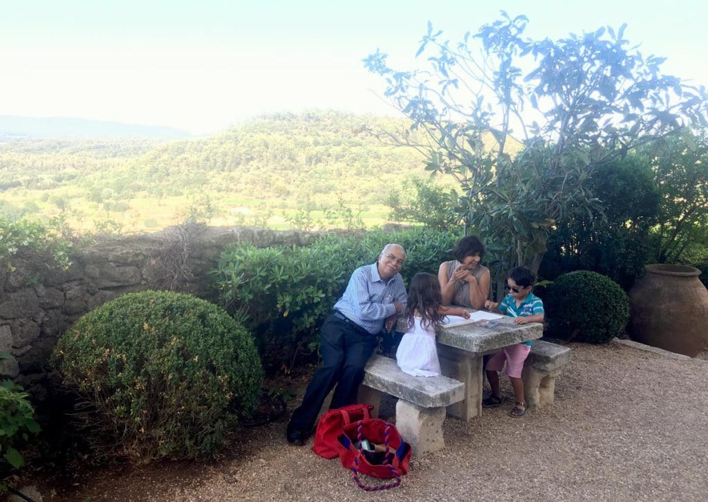 Two kids have lunch with their grandparents on an outdoor table with a view of Menerbes in the distance.