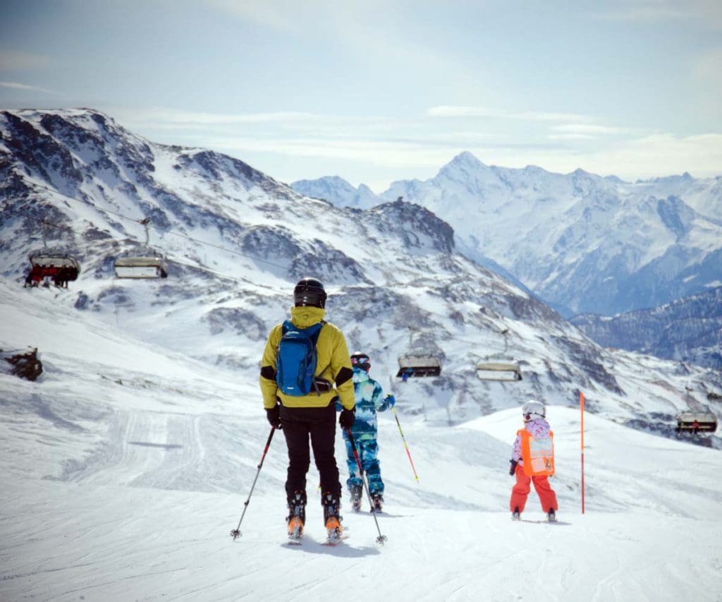 A mom and her two kids ski along a mountain trail in Italy, buying a ski pass can help plan a family ski trip on a budget.