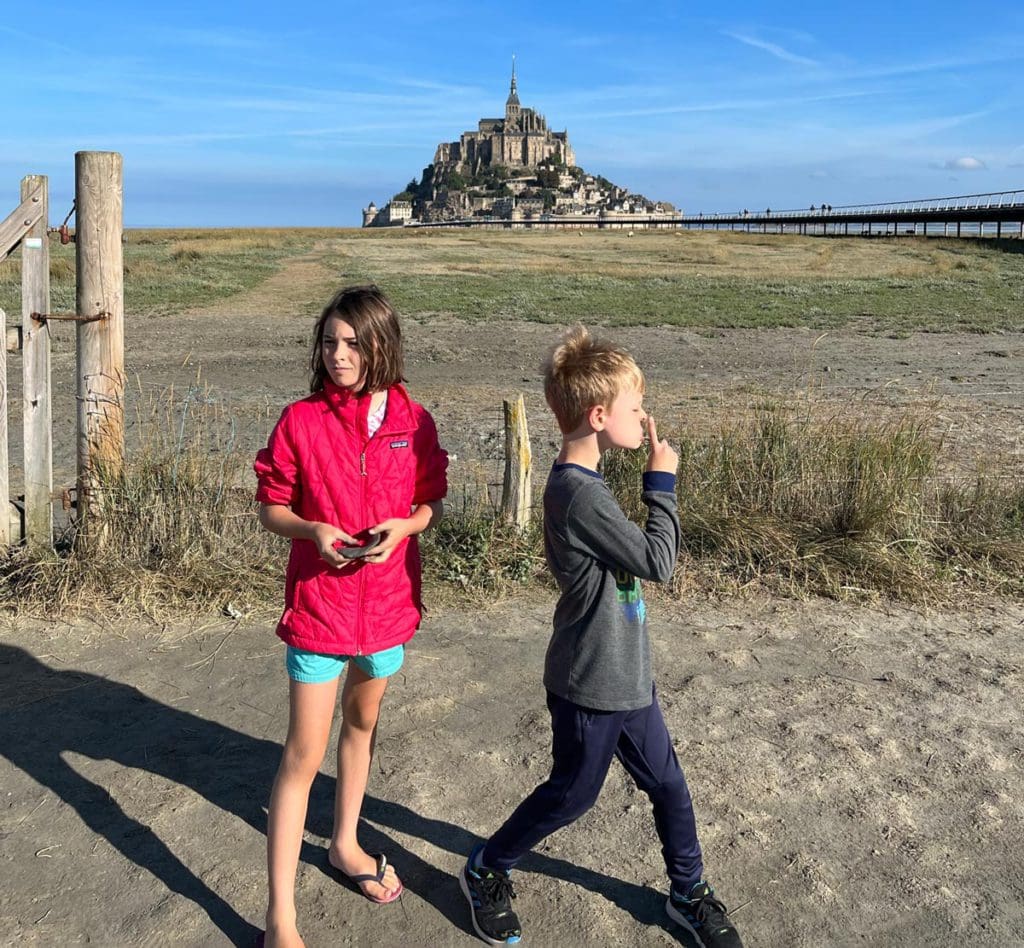 Two kids stand together on a beach, with Mount St. Michel in the distance.