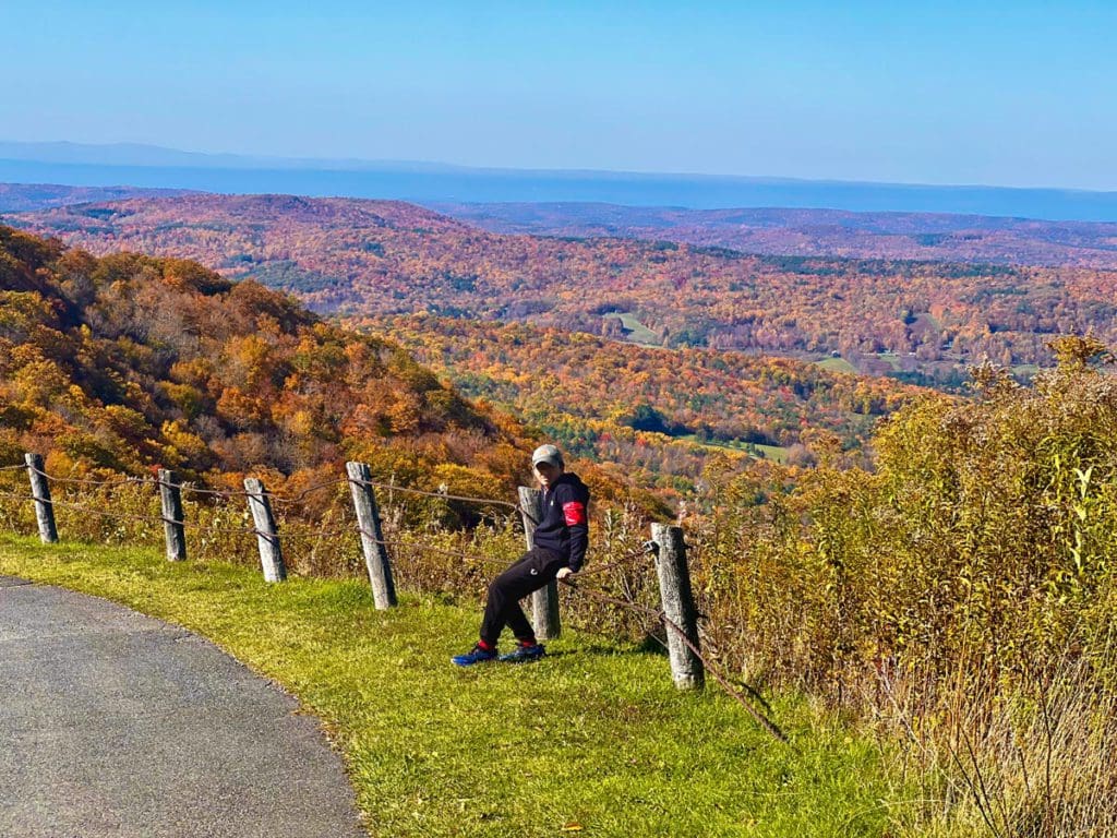 A young boy sits along a fence with an expansive view of colorful fall foliage in the distance at Pittsfield State Forest, one of the best places to visit for fall in New England with kids.