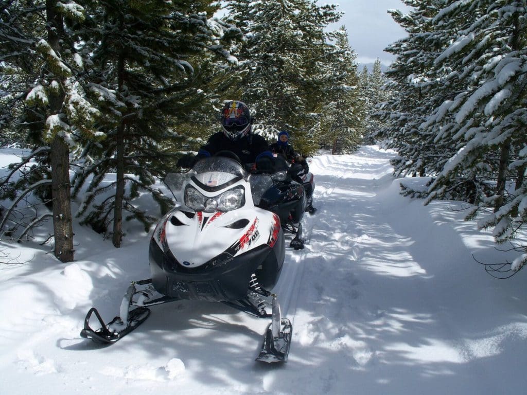 Two snowmobiles travel along a path in Big Sky, with trees flanking each side of the trail.