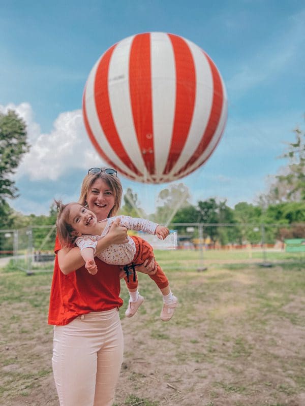 A mom holds her toddler girl, with a large red and white hot air ballon from BalloonFly behind them.