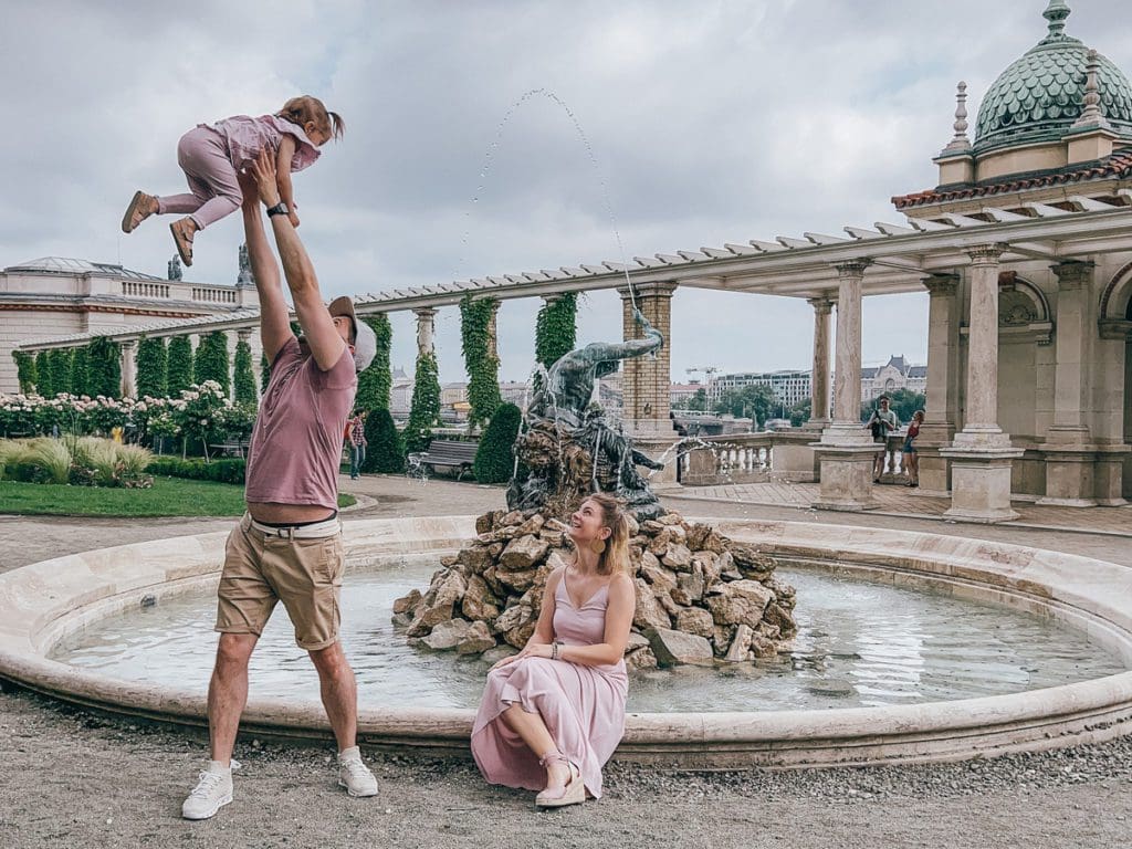 A mom sits along a fountain, while watching dad joyfully throw baby in the air at Castle Garden in Budapest.
