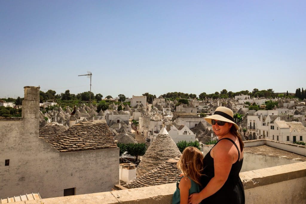A mom and her daughter stand together with a view of Alberobello beyond them.