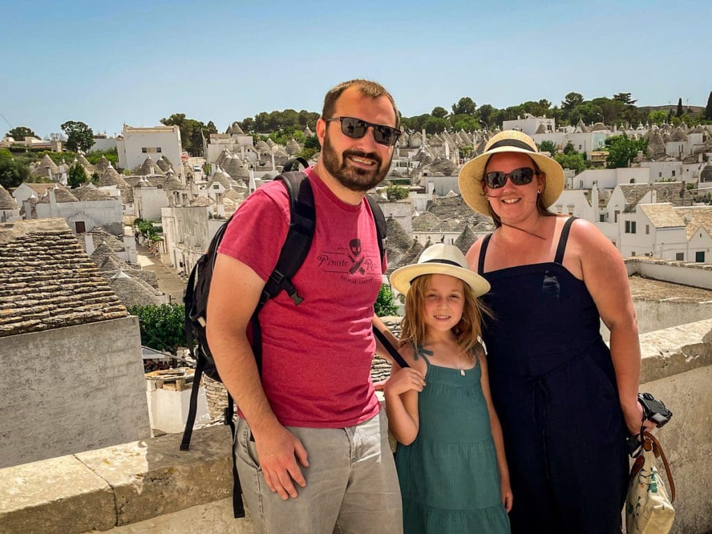 A family of three stands together with the iconic trulli rooftops of Alberobello in the distance, one of the best places to visit in Puglia with kids.