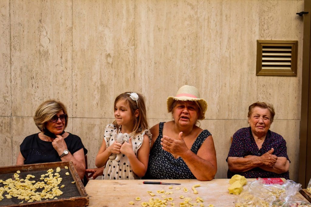 A young girl sits with three Italian women making pasta on a street in Bari, one of the best places to visit in Italy on a budget with kids. 